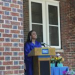 A woman speaking at a podium in front of a brick wall.