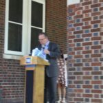 A man standing at a podium in front of a brick building.